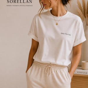 Könnte beinhalten: Eine Frau trägt ein weißes T-Shirt mit hochgekrempelten Ärmeln und dem Text "Dolce Vita Club". Sie trägt auch eine beige Jogginghose. Das Bild enthält den Text "SORELLAN MINIMAL FASHION & LIFESTYLE ESSENTIALS."