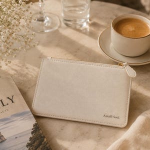 Könnte beinhalten: Eine cremefarbene rechteckige Clutch mit goldenem Reißverschluss und dem Text "Amalfi Soul". Eine Tasse Kaffee auf einer Untertasse, ein Buch über Italien und eine Vase mit Blumen befinden sich ebenfalls auf dem Tisch.