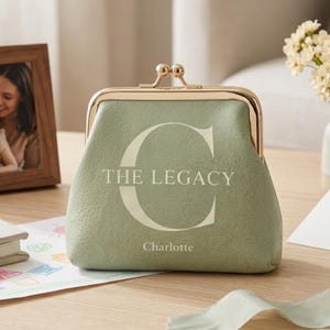 Peut inclure: Porte-monnaie vert sauge avec fermoir et cadre dorés. Le texte "THE LEGACY" et le nom "Charlotte" sont imprimés sur le devant. Une photo encadrée et un petit vase avec des fleurs sont en arrière-plan.