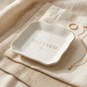 May include: A square white ceramic trinket dish with wavy edges and a gold-colored rim. The center of the dish features the word BLESSED in gold lettering above a small gold cross symbol. A gold ring and a delicate gold chain necklace are nearby.