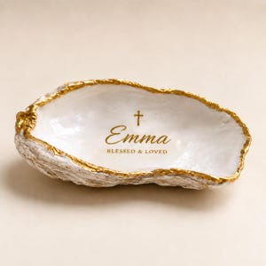 Könnte beinhalten: Eine weiße Austernschalen-Schale mit goldfarbenem Rand. Das Innere zeigt ein goldenes Kreuzsymbol über dem Namen Emma und dem Text BLESSED & LOVED, der in goldener Schreib- und serifenloser Schrift auf die Schale gedruckt ist.