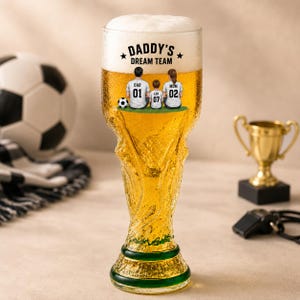 Pode incluir: Uma caneca de cerveja de vidro transparente em forma de troféu, cheia de cerveja dourada e espuma branca. O vidro apresenta um gráfico de três pessoas com camisolas de futebol e o texto DADDY'S DREAM TEAM, DAD 01, LEO 07 e MOM 02.