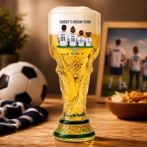 Puede incluir: Un vaso de cerveza alto y transparente con forma de trofeo, lleno de cerveza dorada y espuma. El vaso presenta un gráfico impreso de cuatro personas con camisetas de fútbol con el texto DADDY'S DREAM TEAM, DADDY 01, MOM 10, LEO 07, MIA 11 y EST. 2026.