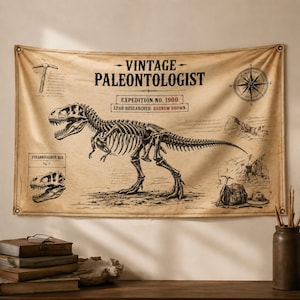 May include: A beige wall tapestry featuring a detailed illustration of a Tyrannosaurus Rex skeleton. The tapestry includes text that reads "VINTAGE PALEONTOLOGIST" and "EXPEDITION NO. 1900". Additional illustrations of tools and a compass are also present.