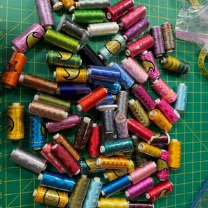May include: A large collection of assorted sewing thread spools in various colors including pink, yellow, blue, green, and metallic tones. Many spools feature labels with the text The Finishing Touch, scattered across a green cutting mat.