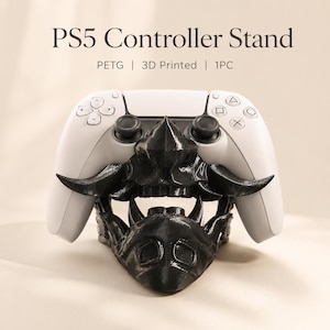 Może przedstawiać: Czarny, wydrukowany w 3D stojak na kontroler z PETG w kształcie stylizowanej maski, podtrzymujący biały kontroler PS5. Tekst PS5 Controller Stand, PETG, 3D Printed, 1PC jest widoczny nad kontrolerem. Stojak posiada ostre, kanciaste detale.