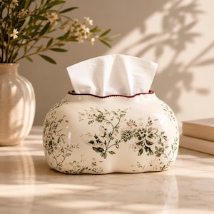 Puede incluir: Una caja para pañuelos de cerámica color crema con forma redondeada y orgánica y un borde superior festoneado. La superficie presenta un detallado patrón floral botánico en verde y un borde fino de color rojo oscuro. Un pañuelo blanco sobresale.