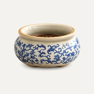 May include: A round ceramic incense burner featuring a cream-colored base with a blue floral vine pattern. The bowl has a smooth, crackled glaze finish and a tan rim. Inside the vessel sits a circular, perforated bronze-toned metal insert.