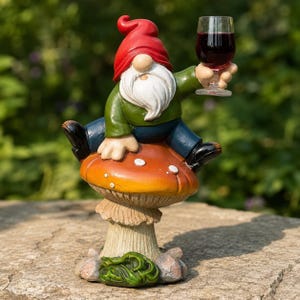Può includere: Una statuetta di gnomo da giardino con cappello rosso, camicia verde e pantaloni blu, seduta sopra un grande fungo arancione e giallo. Lo gnomo ha una lunga barba bianca e tiene in mano un bicchiere trasparente con liquido rosso scuro.