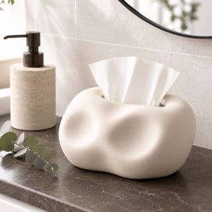 May include: A cream-colored, rounded tissue box cover with an organic, dimpled shape. The smooth, matte-finished container features a central opening at the top from which a single white facial tissue is pulled upward, displaying a soft, folded texture.