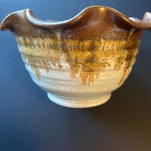 May include: A ceramic bowl with a ruffled, wavy rim. The upper portion features a dark brown glaze that drips down into a textured, speckled light grey and cream base. The exterior surface has horizontal ridges circling the body of the bowl.