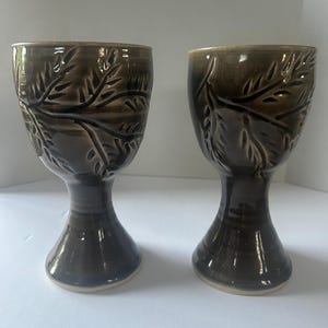 Puede incluir: Dos copas de cerámica hechas a mano con un esmalte marrón oscuro. Cada copa presenta un diseño de hojas y un tallo. Las copas miden aproximadamente 15 cm de altura.