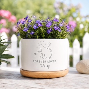 May include: A white ceramic cylindrical planter with a wooden base, filled with purple lobelia flowers. The front features a black line art illustration of a cat reaching for a butterfly, with the text "FOREVER LOVED" and the name "Daisy" below.