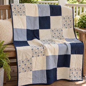 May include: A patchwork quilt featuring a grid of alternating squares in navy blue, light blue, cream, and a white fabric with a small blue floral pattern. The quilt has a dark blue border and is stitched with a diagonal cross-hatch pattern.