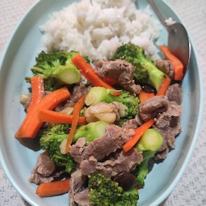 May include: A light blue plate holds a meal of white rice, beef, broccoli, and carrot slices. The beef is brown, the broccoli is green, and the carrots are orange. A silver spoon rests on the plate, ready to eat.