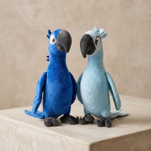 May include: Two plush parrot toys standing side by side. The left parrot is a vibrant royal blue with a dark gray beak and white eye patches. The right parrot is a lighter sky blue with a dark gray beak, white eye patches, and a small crest.