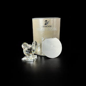 Puede incluir: Una figura de ratón de cristal facetado transparente con ojos negros y bigotes dorados, colocada junto a una caja cilíndrica beige de Swarovski Silver Crystal y un certificado de autenticidad plateado redondo con el logo del cisne.
