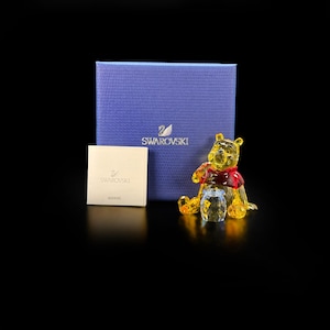 May include: A faceted yellow crystal bear figurine wearing a red crystal shirt, positioned next to a small clear crystal honey pot. Behind the figurine is a blue textured box with the Swarovski logo and a small white booklet with the Swarovski logo.