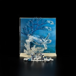Pode incluir: Uma escultura de tartaruga marinha em cristal transparente posicionada em frente a um painel de vidro azul com um ramo de coral azul e dois peixes pequenos. Uma placa de cristal retangular na base exibe o texto Wonders of the Sea by Eternity.