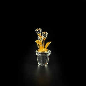 Puede incluir: Una figura decorativa que presenta tres capullos de flor de cristal transparente facetado sobre tallos y hojas de metal dorado, todo ello dentro de una maceta de cristal transparente facetado. La pieza muestra superficies reflectantes.