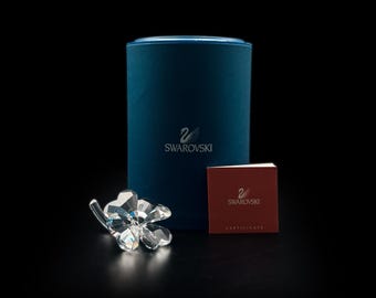 Op de afbeelding: Een helder kristallen beeldje in de vorm van een klavertjevier naast een cilindervormige donkerblauwe Swarovski-doos en een klein vierkant rood certificaatboekje. De doos is voorzien van het zilveren Swarovski-zwaanlogo.