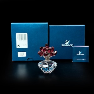 May include: A Swarovski crystal figurine featuring a cluster of deep red rose buds with silver-toned stems emerging from a clear, faceted, diamond-shaped crystal base. The set includes two blue boxes with the Swarovski logo and a certificate booklet.