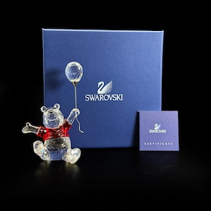 May include: A clear crystal figurine of a bear wearing a red shirt and holding a clear crystal balloon on a thin metal wire. The set includes a blue Swarovski branded box and a matching blue certificate card with the Swarovski logo and text.