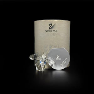 May include: A clear faceted crystal elephant figurine with blue eyes, positioned in front of a cylindrical beige box labeled Swarovski Silver Crystal. A circular silver certificate card with the Swarovski logo and text is placed in front of the box.