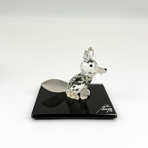 Pode incluir: Uma estatueta de raposa de cristal com um design facetado, sentada sobre uma base retangular preta. A raposa tem olhos pretos e um nariz branco. A base tem uma assinatura em branco. A cauda da raposa é prateada e curvada.