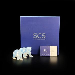 Könnte beinhalten: Zwei schillernde Kristallbärenfiguren vor einer quadratischen blauen Schachtel mit dem silbernen Schriftzug SCS SWAROVSKI CRYSTAL SOCIETY. Neben der Schachtel liegen zwei kleine quadratische Karten, eine davon blau mit dem Text CERTIFICATE und einem Schwanenlogo.