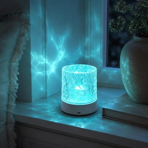 Può includere: Una lampada da tavolo cilindrica con un paralume in vetro cristallino testurizzato che emette una vivace luce turchese. La lampada presenta una solida base circolare bianca con una piccola porta di ricarica visibile sul davanti, creando riflessi.
