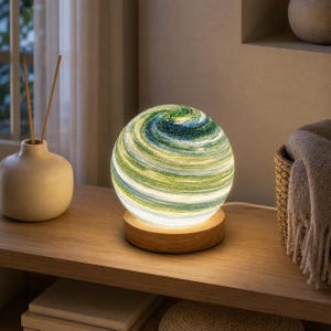 May include: A spherical lamp featuring a swirling pattern of green, blue, and white colours that resemble a planet. The globe rests on a circular light-coloured wooden base, emitting a soft, warm glow from within its textured, translucent surface.