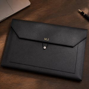 Puede incluir: Una carpeta para documentos de cuero negro con una solapa puntiaguda estilo sobre, un cierre de botón circular plateado con cordón elástico y las iniciales M.J. grabadas en oro en el centro de la solapa frontal del portafolio rectangular.