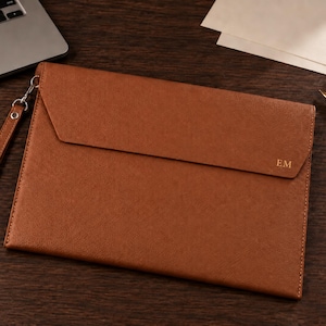 Puede incluir: Un portadocumentos de cuero color canela con una solapa plegable que presenta las iniciales EM grabadas en oro. El estuche rectangular incluye una correa de muñeca a juego desmontable sujeta a un cierre de metal plateado en el lateral.