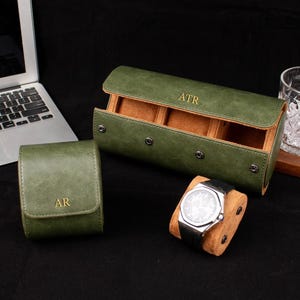 May include: A dark green watch roll with three compartments and gold-colored text reading ATR, alongside a smaller single watch case with the text AR. A wristwatch with a black strap is displayed on a tan cushion in front of the open roll.