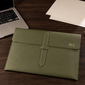 May include: An olive green document sleeve with a textured finish, featuring a vertical strap closure and the gold-embossed initials M.S. on the front flap. The rectangular case is designed to hold papers or a slim device securely.