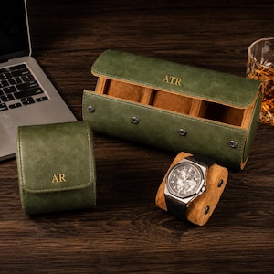May include: Two olive green watch cases with gold monogrammed initials. One case is a single-watch roll with the letters AR, and the other is a three-watch roll with the letters ATR. Both cases feature snap closures and tan interior linings.