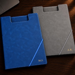 May include: Two rectangular document folders with a cutout top edge and a diagonal elastic strap. One folder is blue with the gold embossed initials M.J., and the other is gray with the gold embossed initials A.R. Both feature a smooth finish.