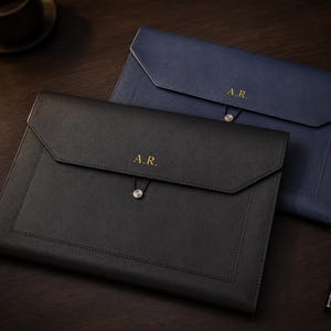May include: A black document folder and a navy blue document folder, both featuring a flap closure with a circular button and elastic cord. Each folder is embossed with the gold-colored initials A.R. on the front flap.