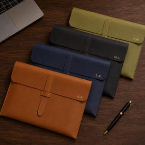 Puede incluir: Cuatro fundas rectangulares para documentos en color tostado, azul marino, negro y verde oliva. Cada funda tiene un cierre de correa vertical e iniciales grabadas en oro: A.R., L.K., M.J. y E.W. Un bolígrafo negro y dorado descansa al lado.