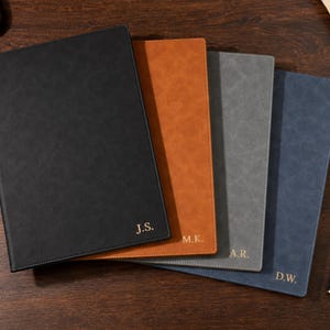 May include: A set of four rectangular notebooks with textured covers in black, tan, grey, and blue. Each notebook features gold-embossed initials in the bottom right corner, specifically J.S., M.K., A.R., and D.W., arranged in a fanned-out stack.