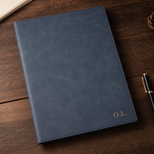 Puede incluir: Un cuaderno rectangular de color azul marino con un acabado mate liso y bordes cosidos. La esquina inferior derecha presenta las iniciales O.L. grabadas en oro. Un bolígrafo negro con detalles dorados se sitúa a la derecha del cuaderno.