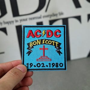 May include: Square patch with a black border and a light blue background. The patch features the text "AC/DC" in red, a lightning bolt, and "BON SCOTT" in yellow. A red cross and the date "19.02.1980" are also present.