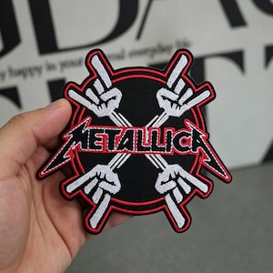 May include: Black and red embroidered patch featuring the word "METALLICA" in red, with white hand gestures and lightning bolts. The patch has a circular shape with a red border and is designed for application on clothing or accessories.
