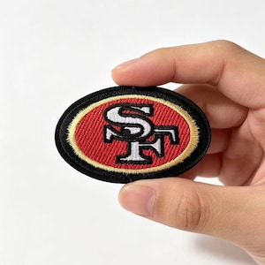 Può includere: Una toppa ricamata di forma ovale con il logo dei San Francisco 49ers. Il design presenta un monogramma SF bianco intrecciato su sfondo rosso, circondato da un sottile bordo dorato e da un bordo esterno spesso di colore nero.