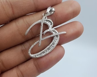 Puede incluir: Un colgante de tono plateado con forma de letra B cursiva, con un acabado metálico e incrustado con numerosas piedras pequeñas, claras y brillantes que cubren toda la superficie de la letra, incluyendo un pequeño lazo en la parte superior.