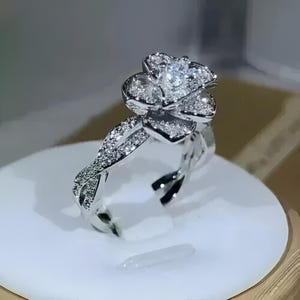 May include: A silver ring featuring a flower design. The flower is made of multiple petals, each encrusted with small, sparkling stones. The band has a twisted design, also adorned with smaller stones. The ring is displayed on a white surface.