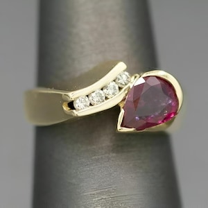 May include: A gold ring featuring a pear-shaped deep red gemstone set in a bezel, accompanied by a curved channel setting containing three small round white diamonds. The band has a smooth, polished finish and a bypass-style design.