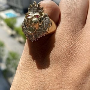 May include: A gold ring featuring a sculpted lion head with a crown, accented by small clear stones set into the mane and two small red stones for the eyes. The lion has an open mouth and is crafted with polished gold details on the face.
