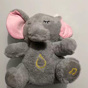 Könnte beinhalten: Ein graues Plüsch-Elefanten-Spielzeug mit rosa Innenohren und geschlossenen Augen. Der Elefant hat eine gelb gestickte Tränenform auf der Brust und eine Musiknote auf dem Fuß. Das Spielzeug ist für Kinder konzipiert.